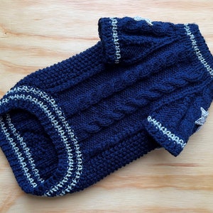 K&ouml;nnte beinhalten: Ein dunkelblauer gestrickter Hundepullover mit wei&szlig;en R&auml;ndern und Stern-Akzenten. Der Pullover hat ein Zopfmuster.
