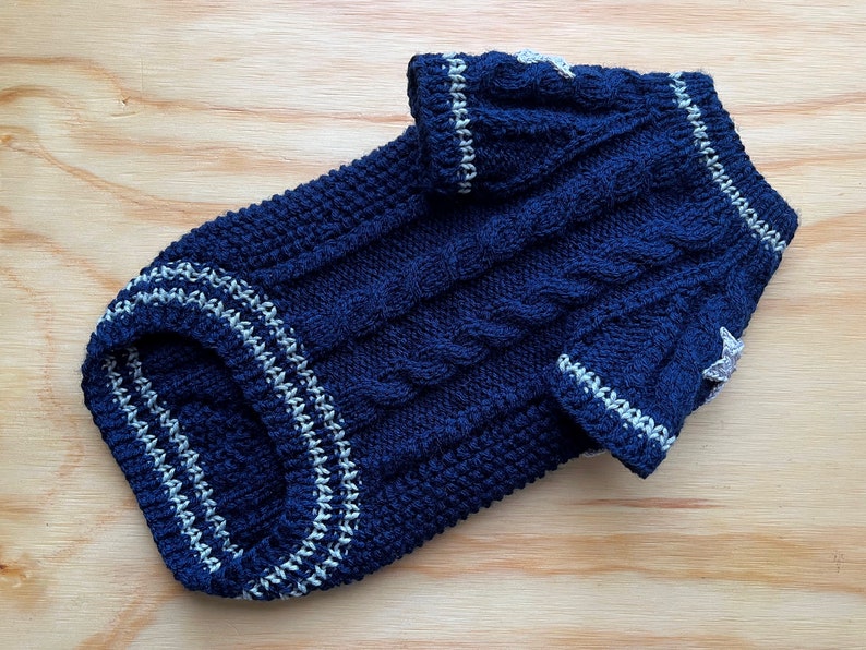 K&ouml;nnte beinhalten: Ein dunkelblauer gestrickter Hundepullover mit wei&szlig;en R&auml;ndern und Stern-Akzenten. Der Pullover hat ein Zopfmuster.