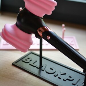 May include: A pink and black plastic hammer with a rose gold accent on the handle. The hammer is on a black stand with the word "BLACKPINK" printed on it.