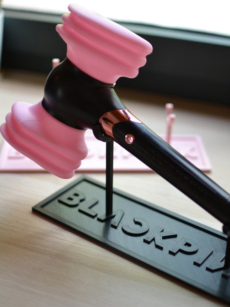 May include: A pink and black plastic hammer with a rose gold accent on the handle. The hammer is on a black stand with the word "BLACKPINK" printed on it.