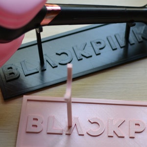 May include: A pink and black 3D printed stand with the word "BLACKPINK" printed on it. The stand is designed to hold a microphone.