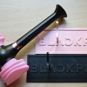 May include: A black and pink BLACKPINK light stick with a microphone design. The light stick has a black handle with the word "BLACKPINK" printed on it. The top of the light stick is pink and shaped like a microphone.