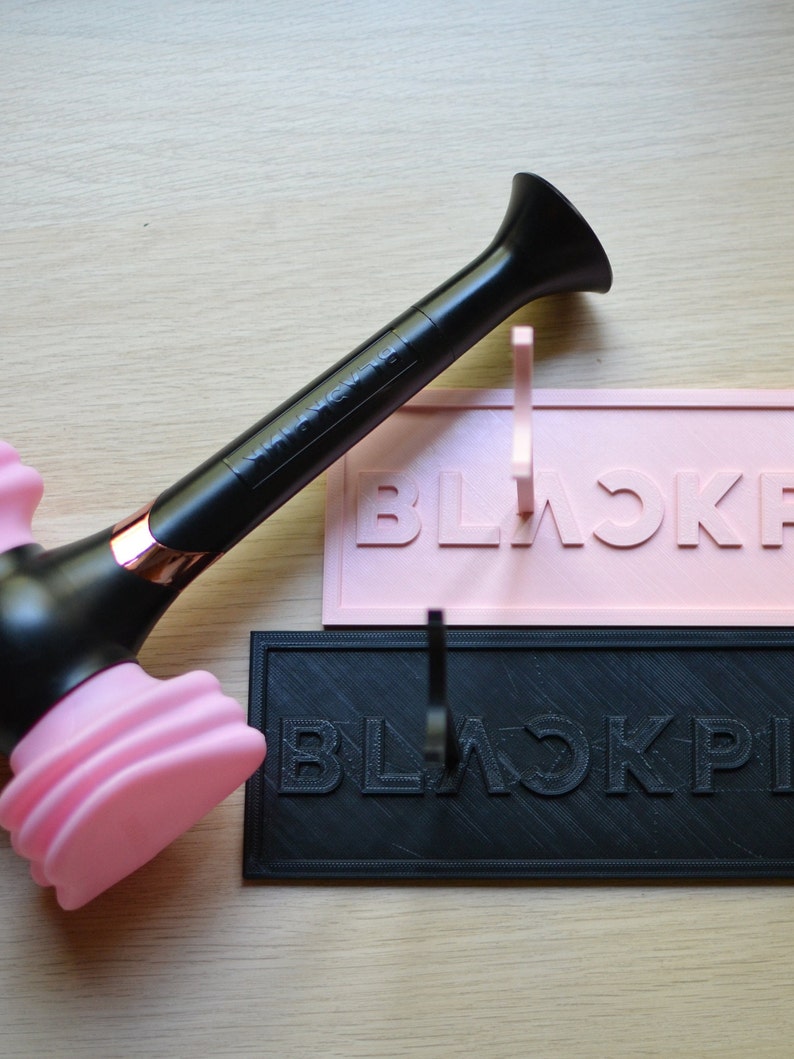 May include: A black and pink BLACKPINK light stick with a microphone design. The light stick has a black handle with the word "BLACKPINK" printed on it. The top of the light stick is pink and shaped like a microphone.