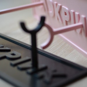 May include: A black and pink 3D printed sign holder with two hooks. The sign holder is made of plastic and has a smooth finish. The sign holder is designed to hold a sign with the word "PINK" printed on it.