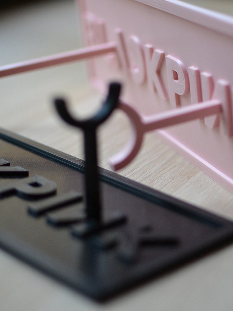 May include: A black and pink 3D printed sign holder with two hooks. The sign holder is made of plastic and has a smooth finish. The sign holder is designed to hold a sign with the word "PINK" printed on it.