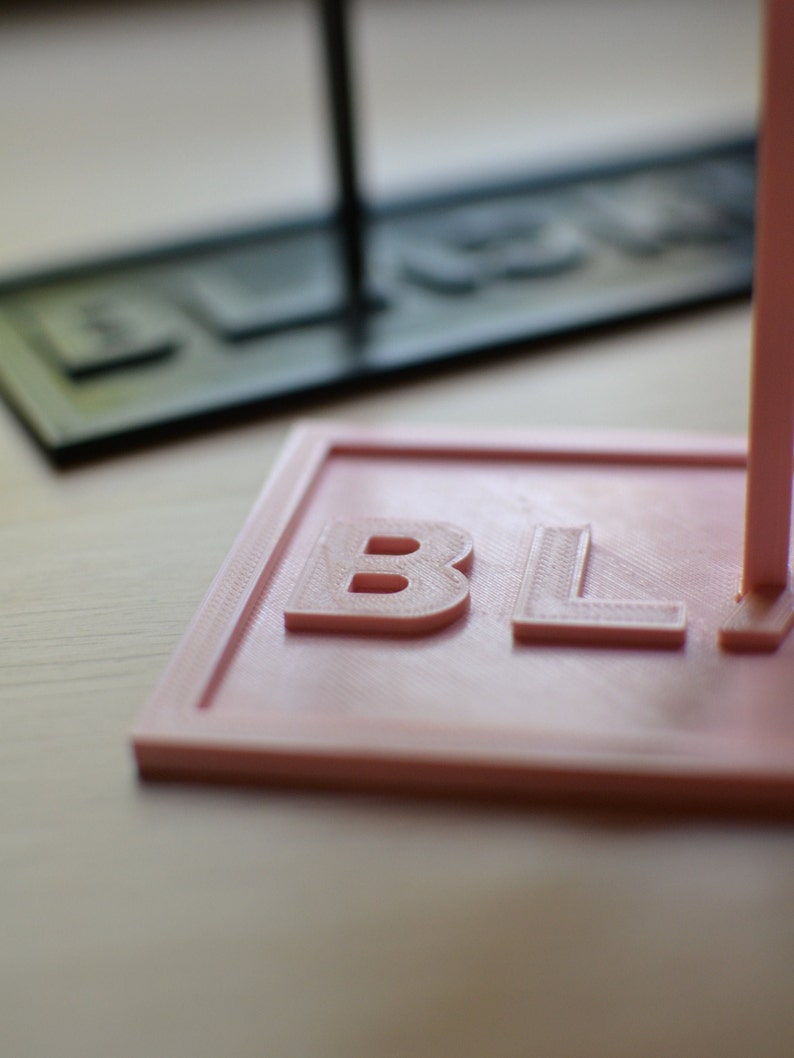 May include: A close-up of a pink 3D printed sign with the word "BLACK" embossed on it.