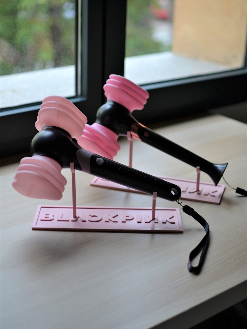 May include: Two pink and black light-up concert hammer-shaped lightsticks with a pink stand that says "BLACKPINK" on it. The lightsticks are on a wooden table.
