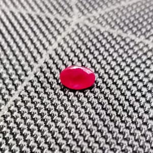 May include: A faceted, oval-shaped, ruby-red gemstone. The gem is set against a textured, gray and white background. The stone's surface reflects light, showcasing its cut and color.