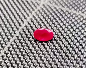 May include: A faceted, oval-shaped, ruby-red gemstone. The gem is set against a textured, gray and white background. The stone's surface reflects light, showcasing its cut and color.