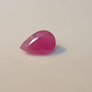 May include: A magenta-coloured, teardrop-shaped gemstone with multiple facets. The polished surfaces of the gem reflect light, highlighting its cut and clarity. The gemstone is set against a neutral background, emphasising its colour and shape.