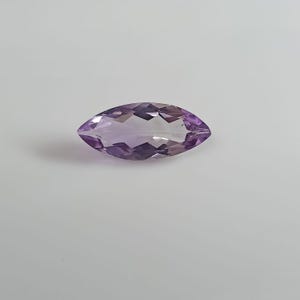 Puede incluir: Una piedra preciosa de amatista con corte marquesa que presenta una superficie facetada con un tono púrpura claro translúcido. La piedra muestra una forma ovalada puntiaguda con cortes geométricos simétricos que reflejan la luz sobre su cuerpo.