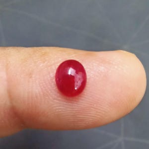 May include: A polished, oval-shaped, deep red gemstone. The gem is smooth and reflective, with a glossy surface. The stone is set against a neutral background, highlighting its rich colour and form. This gemstone could be used in jewellery.