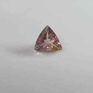 May include: A faceted, triangular-cut gemstone featuring a gradient of soft pink, lavender, and pale yellow hues. The stone displays a clear, translucent quality with sharp, geometric edges and a polished surface that reflects light from its centre.