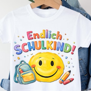 May include: White t-shirt with the German text "Endlich SCHULKIND!" in colorful bubble letters. A large yellow smiley face, a blue backpack, and crayons are also featured. The shirt is hanging on a wooden hanger.