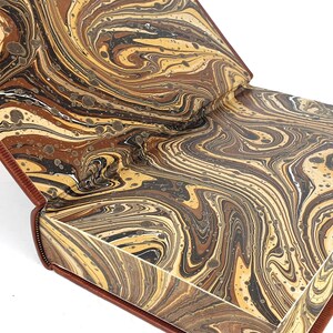 K&ouml;nnte beinhalten: Ein Buch mit einem marmorierten Einband in Braun-, Schwarz- und Cremet&ouml;nen. Das Buch ist ge&ouml;ffnet und zeigt die marmorierten Vors&auml;tze.