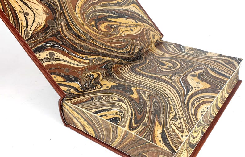 K&ouml;nnte beinhalten: Ein Buch mit einem marmorierten Einband in Braun-, Schwarz- und Cremet&ouml;nen. Das Buch ist ge&ouml;ffnet und zeigt die marmorierten Vors&auml;tze.