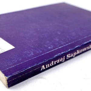 K&ouml;nnte beinhalten: Ein Hardcover-Buch mit lila Einband. Der Name des Autors, Andrzej Sapkowski, ist in Wei&szlig; gedruckt. Auf dem Einband befindet sich ein wei&szlig;es Etikett. Die Seiten sind cremefarben.