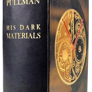 Peut inclure: Un livre relié en cuir noir avec des lettres dorées sur le dos et la couverture. Le titre est "À la croisée des mondes" de Philip Pullman. La couverture présente un cadran d'horloge doré avec des aiguilles rouges.