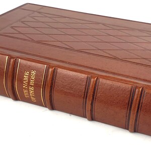 K&ouml;nnte beinhalten: Ein braunes, ledergebundenes Buch mit goldenen Lettern auf dem R&uuml;cken. Der Titel lautet "Der Name der Rose" von Umberto Eco.