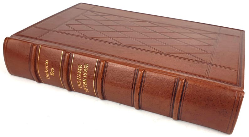 K&ouml;nnte beinhalten: Ein braunes, ledergebundenes Buch mit goldenen Lettern auf dem R&uuml;cken. Der Titel lautet "Der Name der Rose" von Umberto Eco.