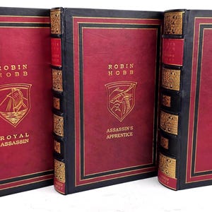 Pode incluir: Três livros de capa dura com capas vermelhas, lombadas pretas e detalhes dourados. Os livros são intitulados "Royal Assassin", "Assassin's Apprentice" e "Assassin's Quest" de Robin Hobb, com emblemas dourados em relevo.