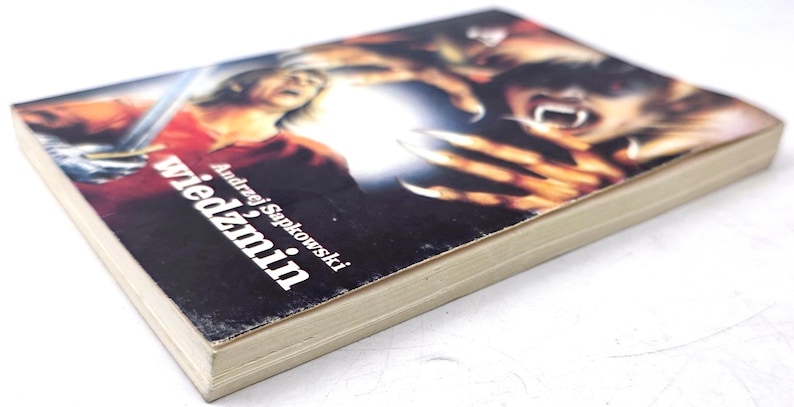 K&ouml;nnte beinhalten: Ein Taschenbuch mit dem Titel "wiedzmin" von Andrzej Sapkowski. Das Cover zeigt eine dramatische Illustration mit einer Figur, die ein Schwert h&auml;lt, und einem Monster mit scharfen Krallen und Z&auml;hnen. Das Buch hat ein abgenutztes Aussehen.
