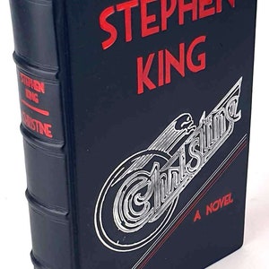 May include: A blue hardcover book with red lettering on the spine and front cover. The title is "Christine" by Stephen King. The front cover features a silver illustration of a car with the word "Christine" in a stylized font.