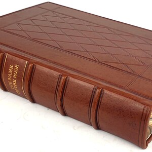 K&ouml;nnte beinhalten: Ein braunes, ledergebundenes Buch mit goldenen Lettern auf dem R&uuml;cken. Der Titel lautet "Der Name der Rose" von Umberto Eco.