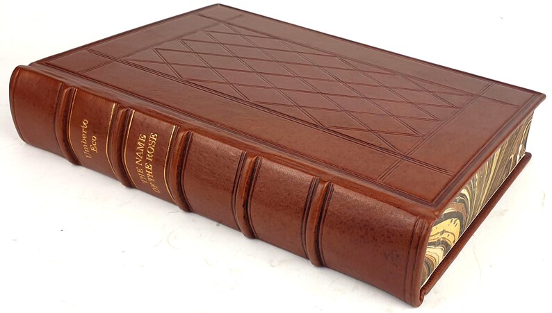 K&ouml;nnte beinhalten: Ein braunes, ledergebundenes Buch mit goldenen Lettern auf dem R&uuml;cken. Der Titel lautet "Der Name der Rose" von Umberto Eco.