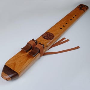 May include: A wooden Native American flute with a brown leather strap and a carved design of a tree and a circle with a face. The flute has six holes and is made of cherry wood.