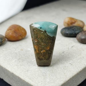May include: A polished gemstone with a trapezoid shape. The top portion is a vibrant turquoise color, transitioning to a textured, earthy brown and green lower section. The stone is displayed on a light gray surface.