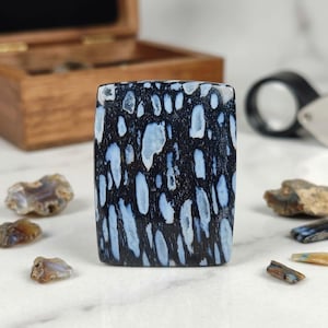 May include: A rectangular, polished gemstone with a dark blue base and irregular, light blue inclusions. The stone is surrounded by smaller, rough-cut gemstones and sits on a white surface. A wooden box and magnifying glass are in the background.
