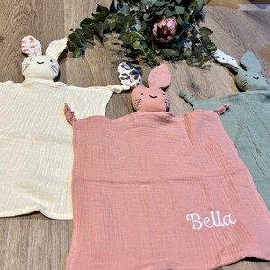 Puede incluir: Tres mantas de seguridad de peluche suave en forma de conejo, en blanco, rosa y verde. La manta rosa tiene el nombre "Bella" bordado.