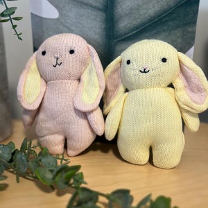 May include: Two knitted plush rabbit toys, one in soft pink with yellow inner ears and the other in pale yellow with pink inner ears. Both rabbits feature black embroidered eyes, a small pink stitched nose, and long floppy ears.