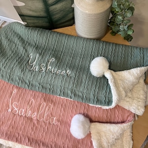 May include: Two knitted baby blankets with pom-poms. The top blanket is green with the name "Yashveer" embroidered on it. The bottom blanket is pink with the name "Isabella" embroidered on it.