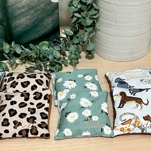 May include: Eight small, square, fabric-covered heat packs with different patterns. The patterns include a leopard print, a floral print, a woodland animal print, and a rainbow print. Each heat pack has a name embroidered on it.