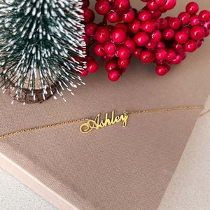 May include: A gold-colored necklace with the name "Ashley" in a cursive font. The necklace is displayed on a neutral-toned surface, with a small decorative Christmas tree and red berries in the background.