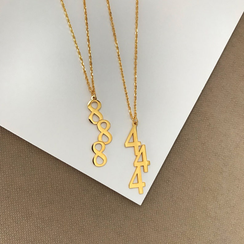 May include: Two gold necklaces with pendant charms. One charm is a vertical stack of four number eight charms. The other charm is a vertical stack of four number four charms.
