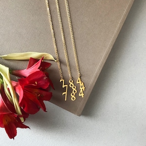 May include: Four gold necklaces with numbers 77, 88, 44, and 4 hanging from them. The necklaces are on a brown surface with red flowers.