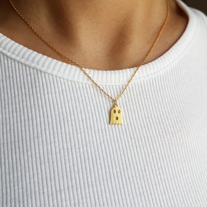 May include: A gold chain necklace with a small ghost pendant. The ghost has two eyes and a mouth.