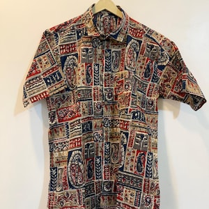 May include: Short-sleeved button-down shirt with a colourful paisley and geometric pattern in red, navy blue, tan, and grey. The shirt is hanging on a wooden hanger against a white background. The shirt has a classic collar.