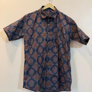 Op de afbeelding: Een overhemd met korte mouwen met een marineblauwe achtergrond en een herhalend paisley-patroon in rood en wit. Het overhemd hangt aan een houten hanger tegen een effen achtergrond.