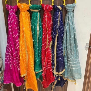 May include: A collection of six colorful scarves hanging on a yellow hanger. The scarves are in shades of pink, orange, green, red, blue, and teal. Each scarf has a patterned design and gold trim.