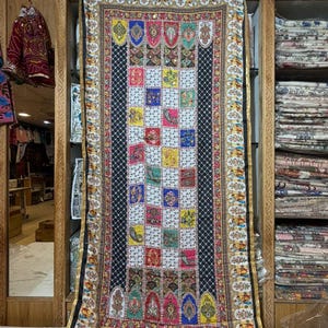 May include: A rectangular textile with a black and white checkered center, framed by ornate borders. The center features colorful squares with various patterns. The borders have a gold trim and detailed designs. The textile is displayed in a wooden cabinet.