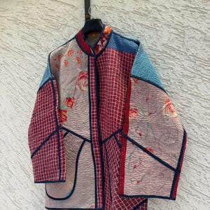 Könnte beinhalten: Eine gesteppte Jacke mit einem Patchwork-Design aus hellrosa Stoff mit roten Blumenmustern, dunkelrotem Stoff mit weißem Gittermuster und einfarbigem blauem Stoff. Die Jacke hat einen Stehkragen, lange Ärmel und eine dunkelblaue Paspelierung.