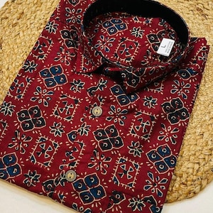 May include: A maroon button-down shirt with a black collar and a repeating pattern of blue and black shapes. The shirt has wooden buttons and is made of a soft fabric. The shirt is a size large.