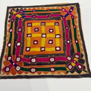 Op de afbeelding: Een vierkant, geborduurd textiel met een centraal oranje en geel patroon. Het ontwerp heeft een raster van kleine vierkantjes en wordt omlijst door een rand van roze, groen en oranje. Kleine, ronde spiegels zijn in het ontwerp verwerkt.