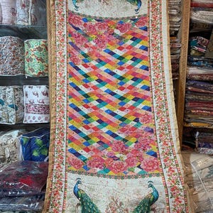 Peut inclure: Textile coloré avec un motif géométrique multicolore. Le tissu présente un fond crème avec des motifs floraux et de paons. Le motif central est un motif géométrique vibrant et multicolore, encadré de motifs floraux et de paons.