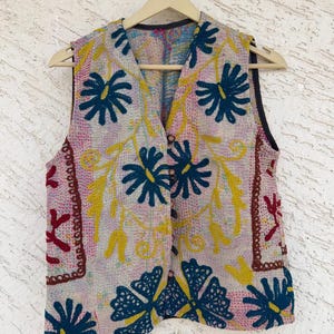 Puede incluir: Un chaleco sin mangas con cuello en V, con un diseño floral. El chaleco es principalmente rosa claro con flores bordadas en azul oscuro y amarillo. Tiene un ribete marrón oscuro y está colgado de una percha de madera.