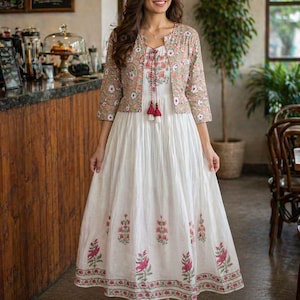 May include: A white maxi dress with floral embroidery and a matching floral print jacket. The dress features a round neckline and a flowing skirt. The jacket has three-quarter sleeves. The dress is adorned with pink and green floral patterns.